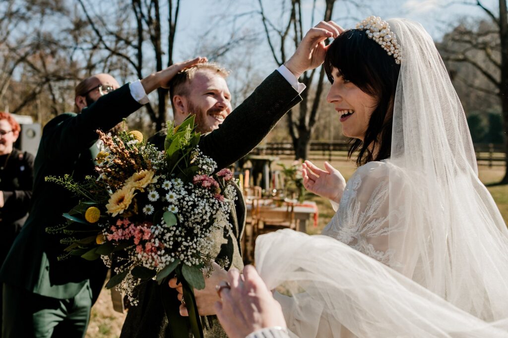 A bride in a white veil holding a bouquet of flowers smiles as a groom and guests joyfully pat each other’s heads during an outdoor wedding ceremony, beautifully captured by a Nashville wedding photographer on a sunny day.