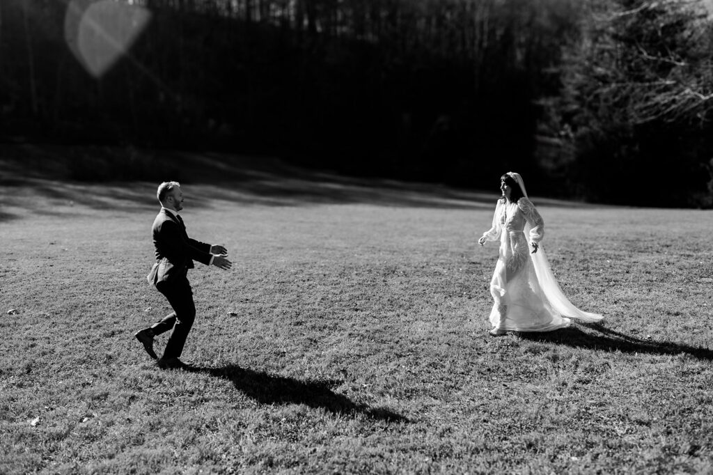 A groom in a suit runs toward a bride in a white dress on a grassy field, both smiling, with trees and sunlight behind them. Captured by a Nashville wedding photographer, the black and white image with blurred edges highlights their joyful moment.