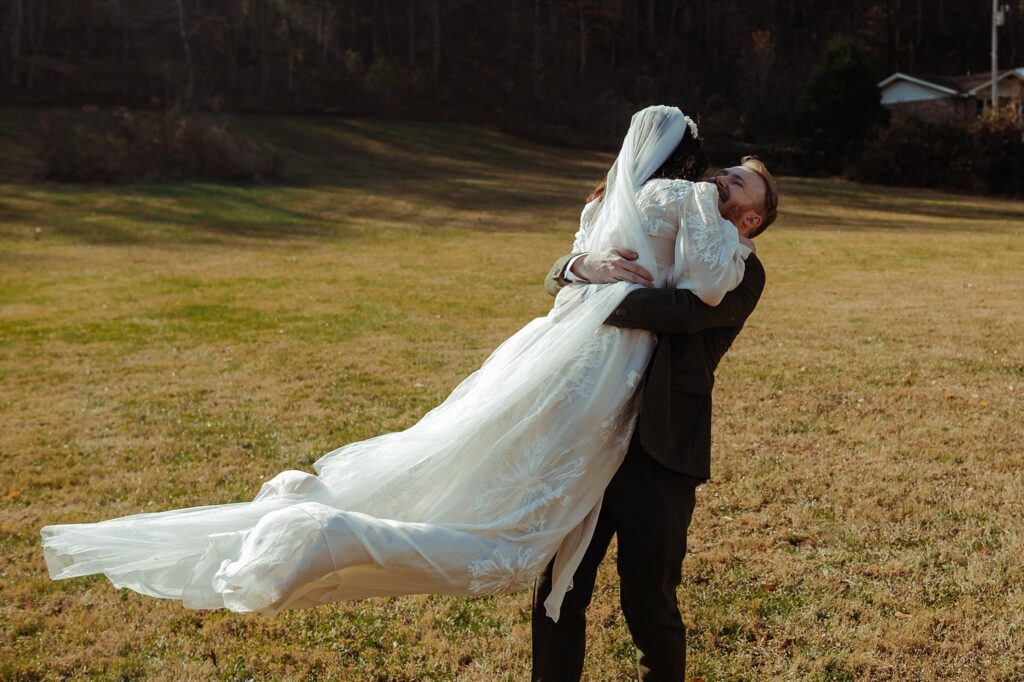 A groom joyfully lifts his bride, who is wearing a long white wedding dress and veil, in a grassy outdoor field as captured by a Nashville wedding photographer, with trees and a house in the background.