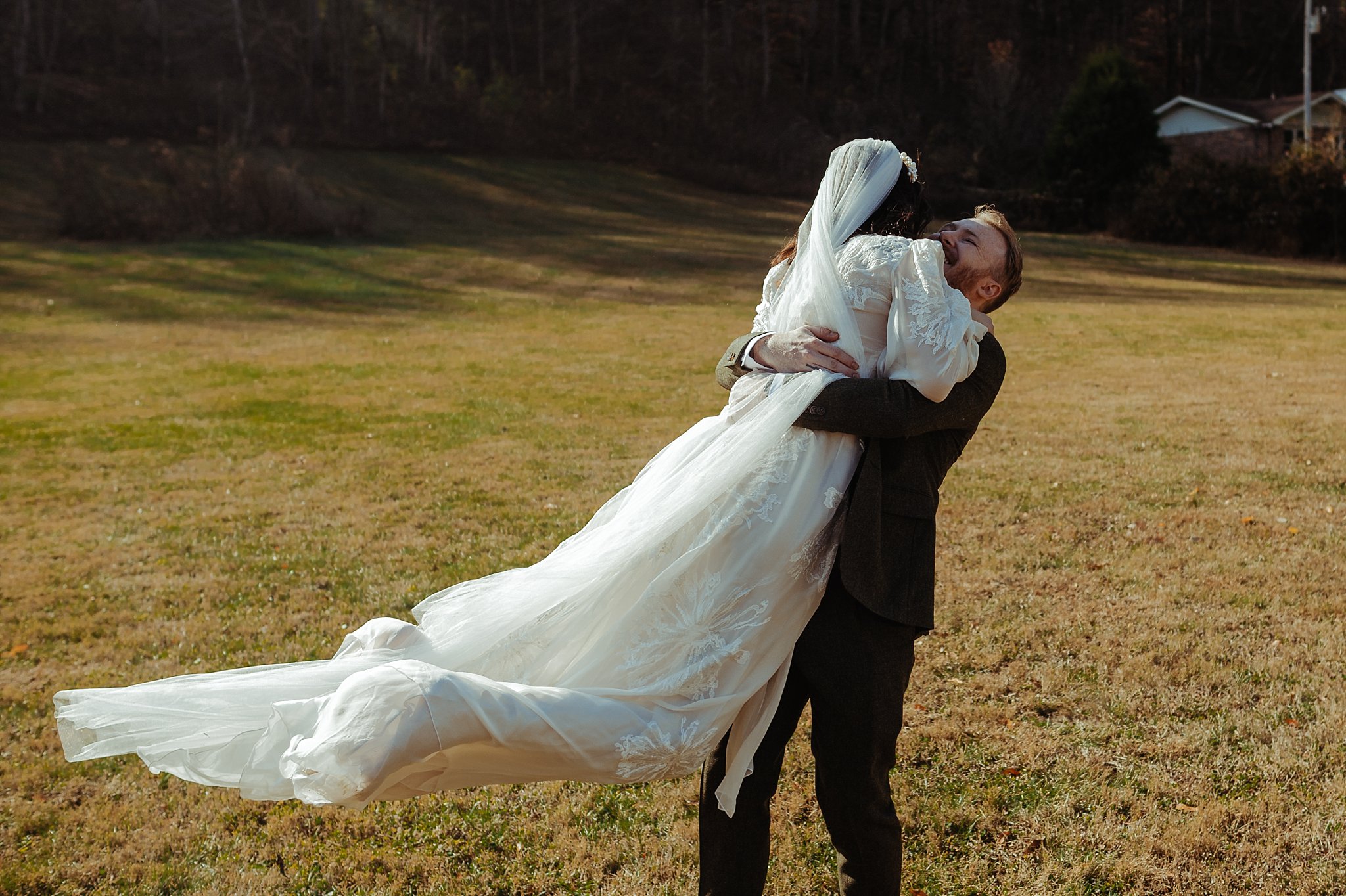 A groom joyfully lifts his bride, who is wearing a long white wedding dress and veil, in a grassy outdoor field as captured by a Nashville wedding photographer, with trees and a house in the background.
