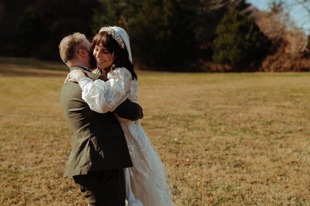 A groom in a suit lifts and embraces a smiling bride in a white wedding dress as they celebrate outdoors on a grassy field—captured perfectly by a Nashville wedding photographer with trees in the background.