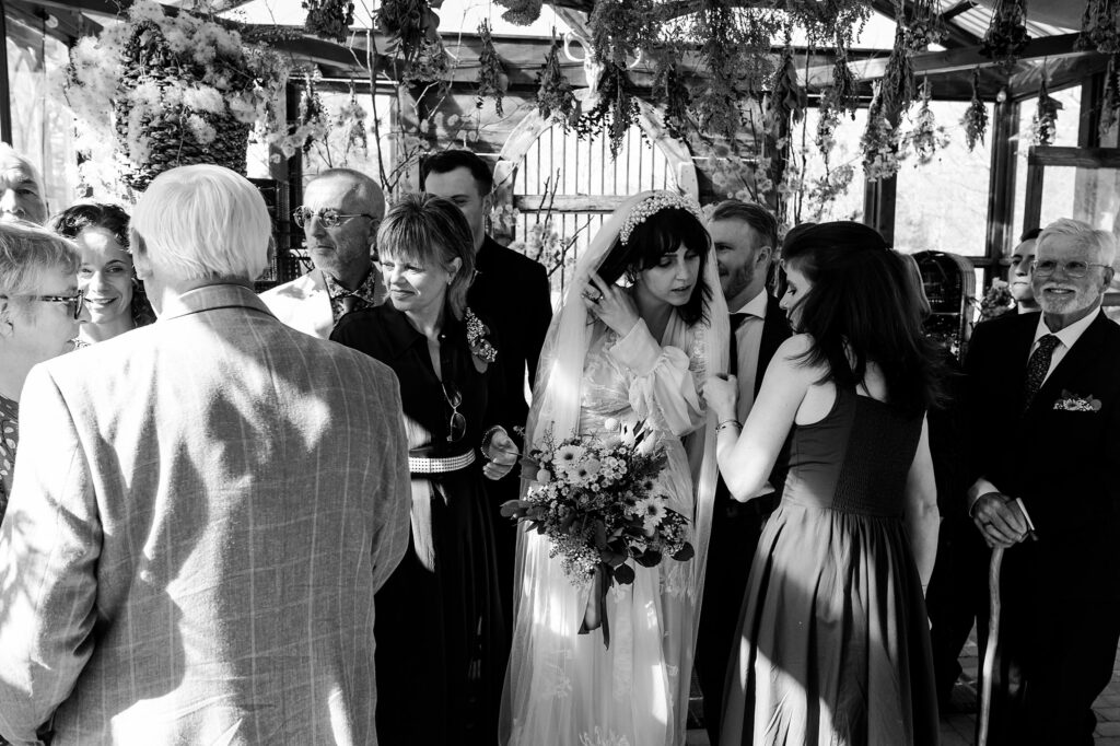 A bride holding a bouquet stands among a group of smiling guests at an outdoor wedding, with plants hanging overhead and sunlight streaming through the structure, perfectly captured by a Nashville wedding photographer.