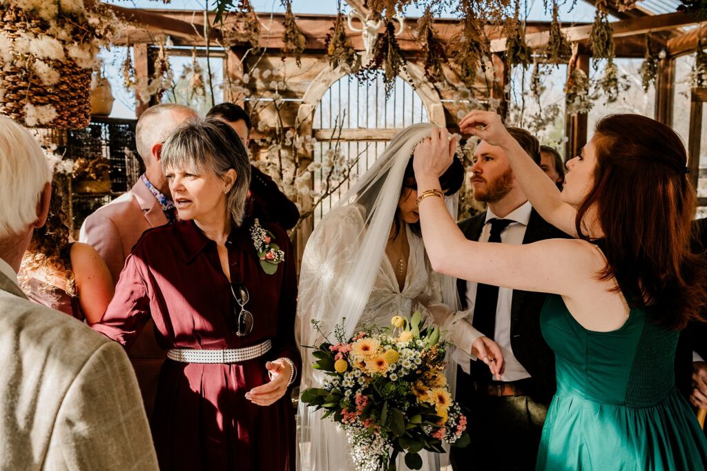 A bride holding a colorful bouquet stands surrounded by people as a woman in a green dress adjusts her veil. Sunlight streams through the rustic, plant-filled setting, captured beautifully by a Nashville wedding photographer. Others watch in formal attire.