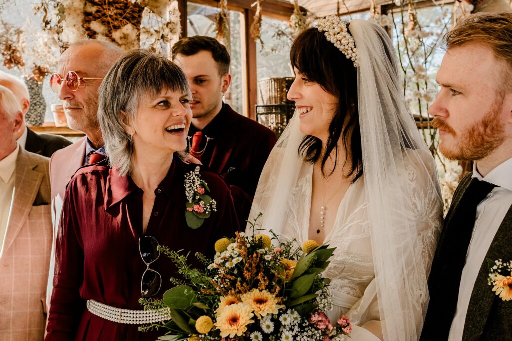 A smiling bride in a veil and holding a bouquet stands with a happy group of people indoors, including a woman with gray hair, as sunlight streams through the windows—captured beautifully by a Nashville wedding photographer.