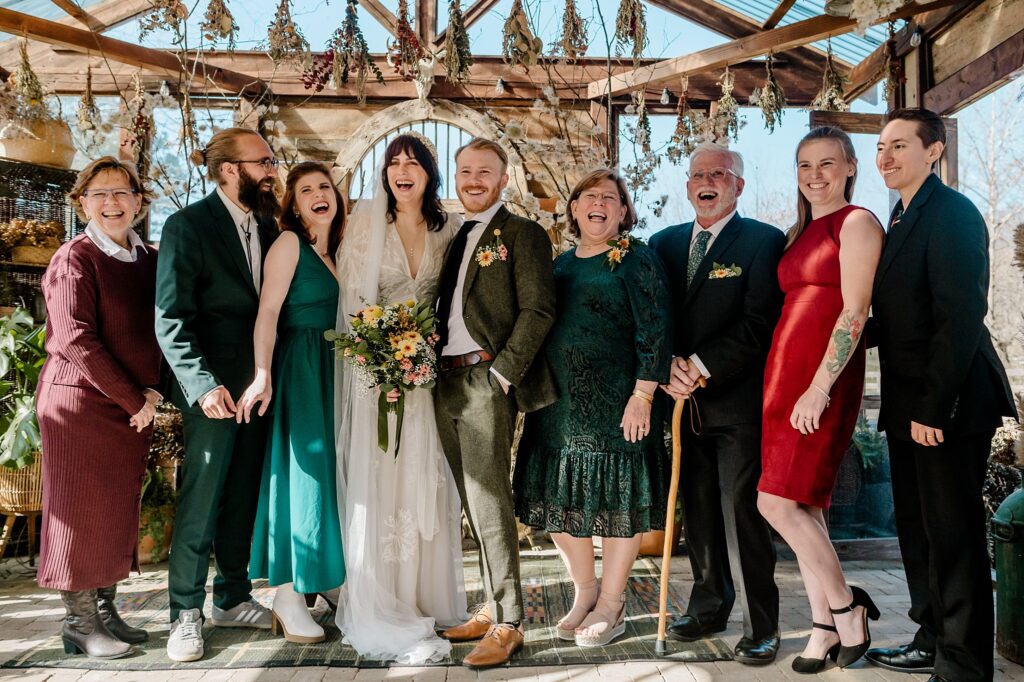 A joyful wedding group poses together indoors under a glass roof, laughing and smiling. Captured by a Nashville wedding photographer, the bride, groom, and party wear colorful attire, surrounded by dried flowers and sunlight.