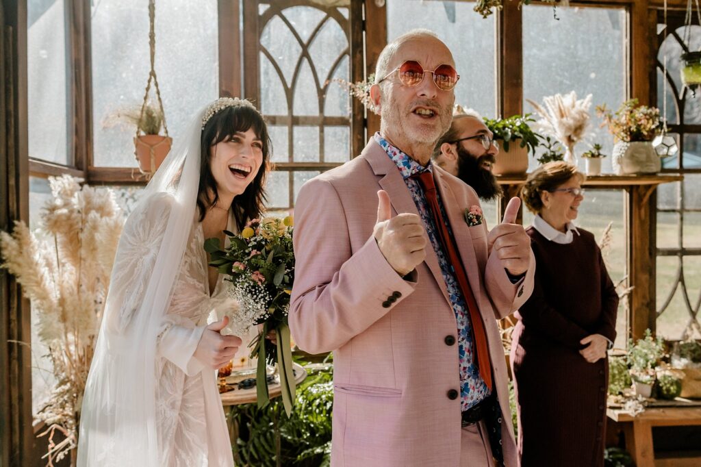 A bride in a white dress laughs while an older man in a pink suit and pink sunglasses gives two thumbs up. Captured by a Nashville wedding photographer, the group smiles in a sunlit, plant-filled room filled with joy.
