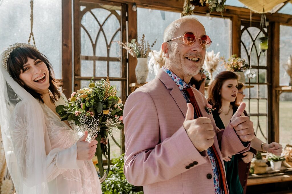 A smiling bride in a white dress stands next to an older man in a pink suit and sunglasses giving two thumbs up, captured perfectly by a Nashville wedding photographer, with a bridesmaid and floral decorations in the background.