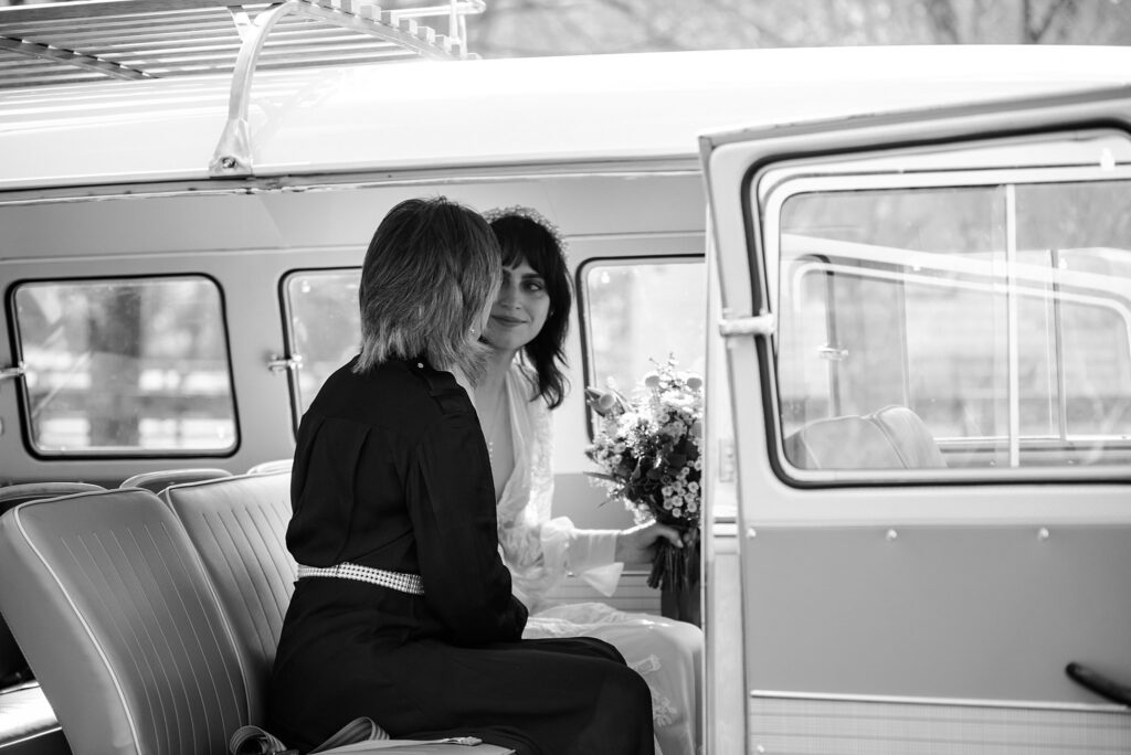 Two women sit facing each other in the back of a vintage van, one in white holding a bouquet, the other in dark attire. Captured in timeless black and white by a Nashville wedding photographer, the van door stands open to the world.