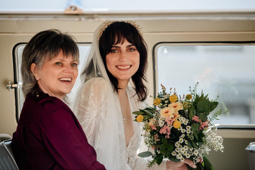 A bride in a white dress and veil, holding a bouquet of flowers, sits next to an older woman in maroon. Both are smiling inside a sunlit vehicle—captured beautifully by a Nashville wedding photographer.