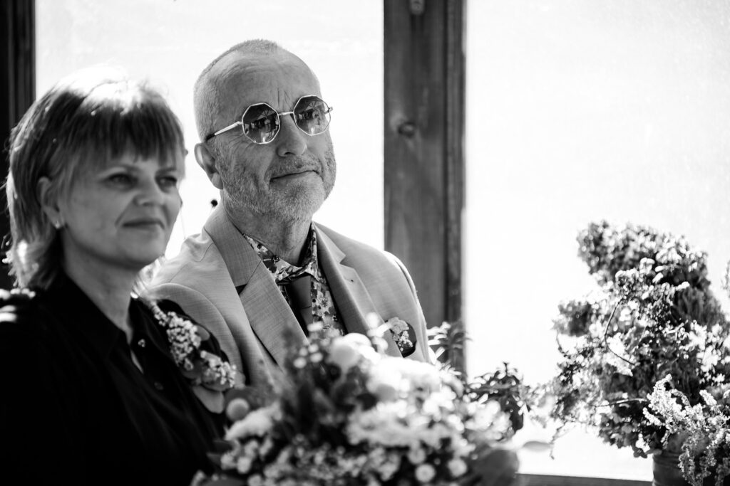 A black and white photo by a Nashville wedding photographer captures an older man in round sunglasses and a suit, sitting beside a woman. Both hold floral bouquets, likely at a wedding, as light streams in from behind them.