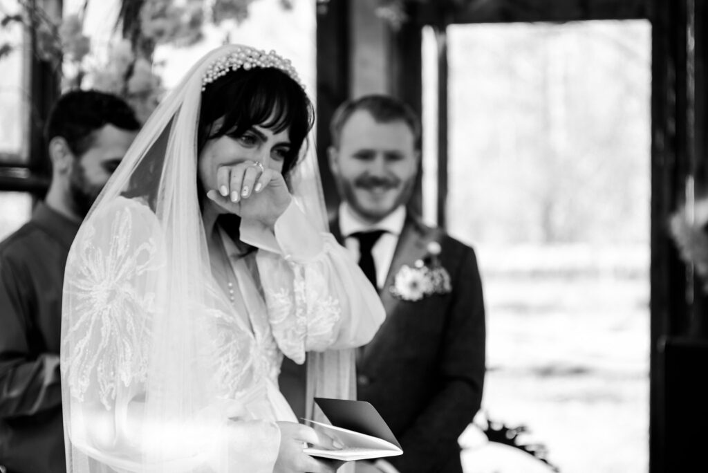 A bride in a veil wipes away tears while holding a booklet, as a smiling groom and another man stand behind her at the ceremony. Captured in black and white by a Nashville wedding photographer.