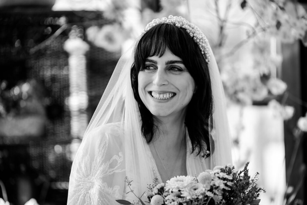 A bride with dark hair, wearing a pearl headband and veil, smiles while holding a bouquet of flowers. Captured by a Nashville wedding photographer, the softly blurred background highlights beautiful floral decorations.