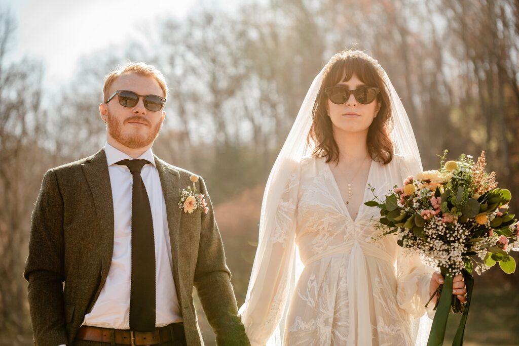 A bride and groom wearing sunglasses hold hands outdoors as their nashville wedding photographer captures the moment. The bride wears a white dress and veil, holding a bouquet, while the groom dons a suit with tie and boutonniere against blurred trees.