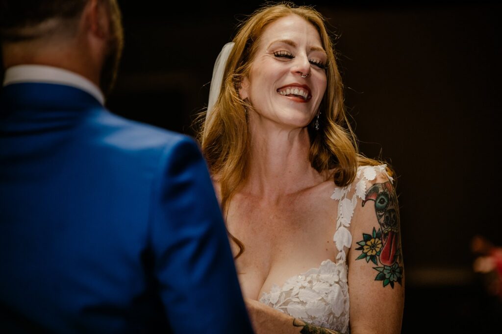 A joyful bride with long red hair and a veil smiles brightly during a wedding ceremony, wearing a lace dress and showing colorful tattoos on her arm, perfectly captured by a Nashville wedding photographer. A person in a blue suit stands beside her, back to the camera.