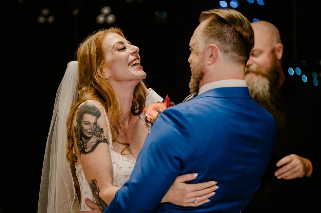 A bride in a white dress and veil smiles joyfully at her groom in a blue suit as they embrace, with a bearded officiant behind them. Captured by a Nashville wedding photographer, the bride’s large arm tattoo stands out against the dark, blurred background.