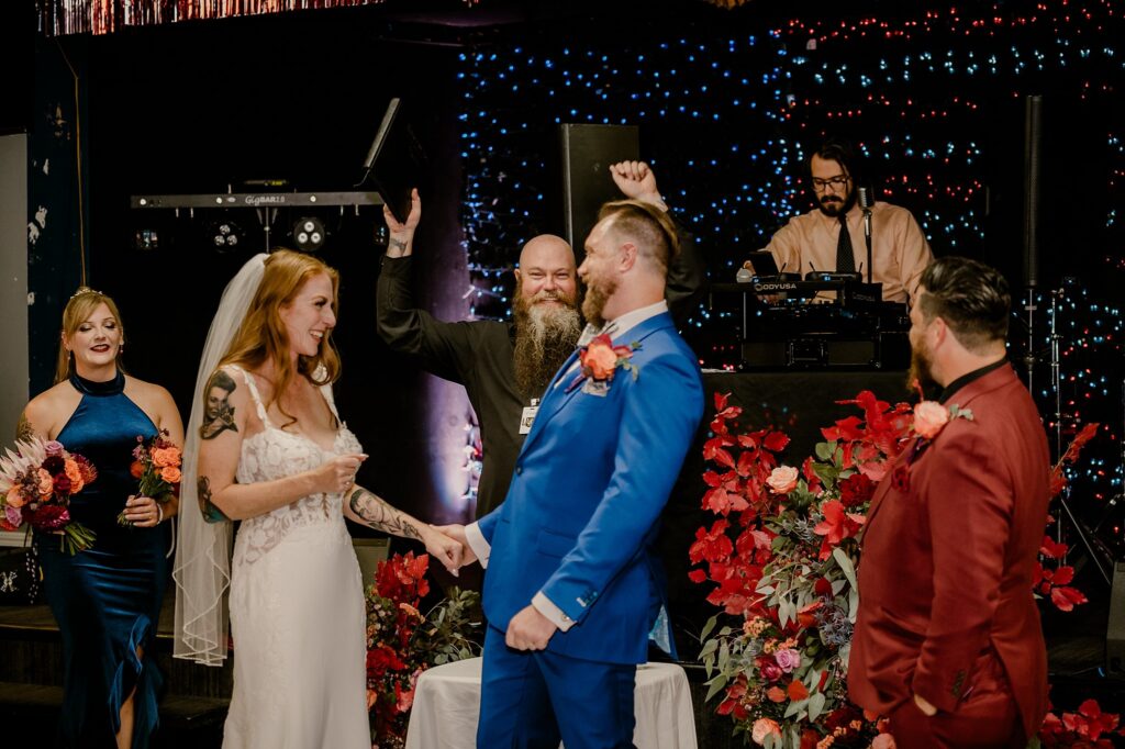 A bride and groom smile and hold hands at their wedding ceremony, as a Nashville wedding photographer captures the moment. Friends with flowers stand nearby, a DJ raises his hand, and red floral decorations surround them.