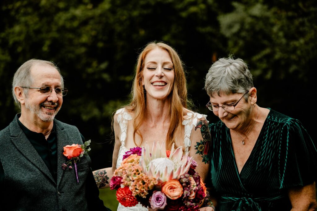 A smiling bride holding a colorful bouquet stands between two older adults, all dressed in formal wear, outdoors with greenery in the background, beautifully captured by a Nashville wedding photographer.