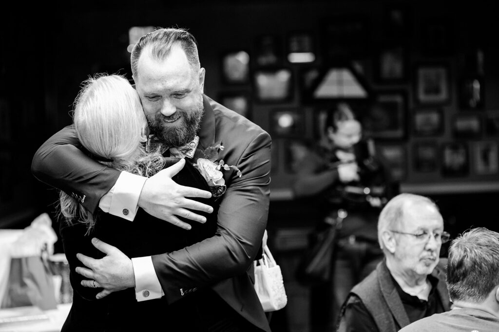 A bearded man in a suit warmly hugs a woman with light hair at an indoor event. Captured by a Nashville wedding photographer, the joyful, candid scene features guests seated in the background and framed photos decorating the wall.