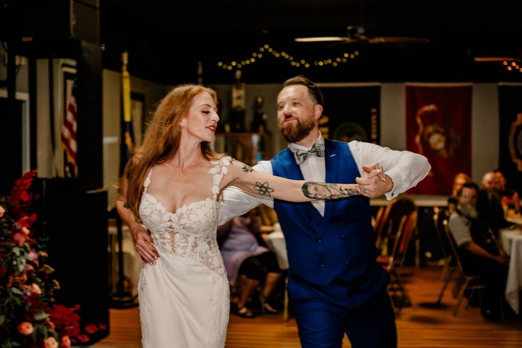 A bride in a white lace dress and a groom in a blue suit dance together indoors, smiling and holding hands. Captured by a Nashville wedding photographer, guests sit at tables under soft lighting and hanging string lights.