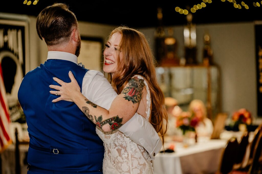 A bride in a lace dress with visible tattoos smiles while dancing with a man in a blue vest, beautifully captured by a Nashville wedding photographer. Guests are seated at decorated tables in the background.