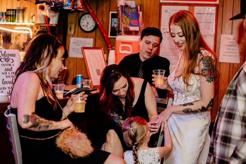 A bride in a white dress smiles while a young girl touches her gown, surrounded by three other adults with drinks, chatting and laughing in a cozy, warmly lit bar—captured perfectly by a Nashville wedding photographer.