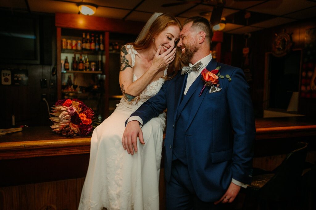 A bride in a white dress and veil sits on a bar counter, laughing with her bearded groom in a blue suit and bow tie. Captured by a Nashville wedding photographer, their joy shines as she holds vibrant flowers in the warmly lit bar.