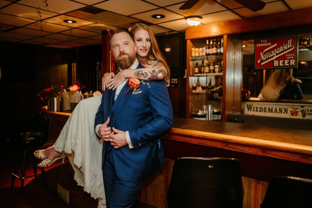 A bride in a white dress playfully sits on a bar, embracing her bearded groom in a blue suit. Captured by a Nashville wedding photographer, they smile warmly amid vintage beer signs and bottles in this cozy bar setting.