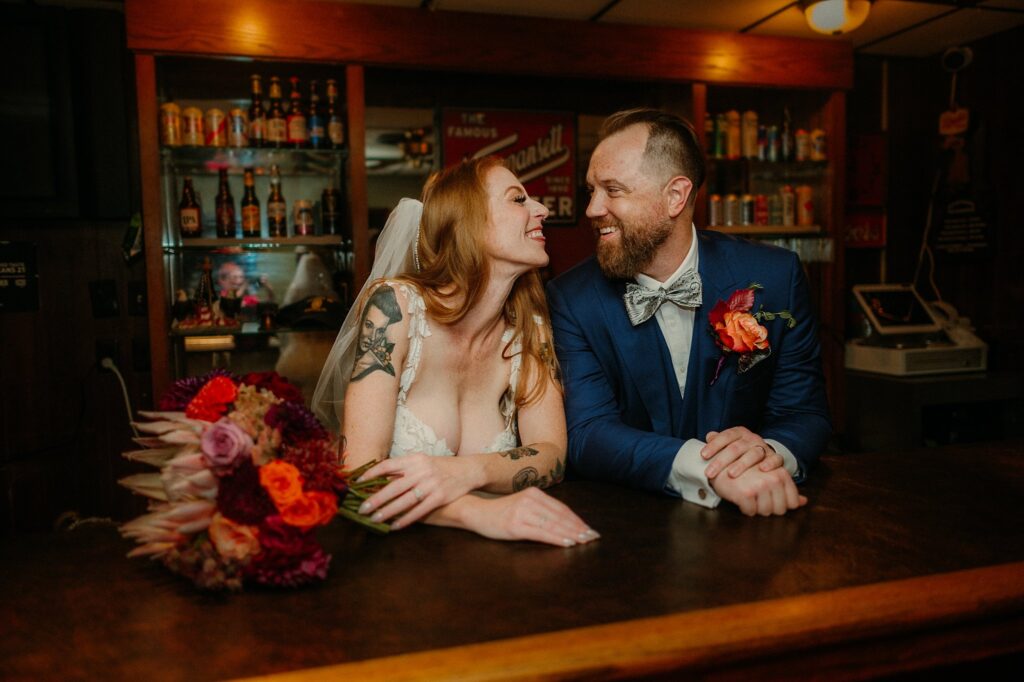 A bride and groom in wedding attire lean on a bar counter, smiling and gazing at each other. Captured by a Nashville wedding photographer, the bride holds a bouquet of colorful flowers, with bottles and snacks visible in the background.
