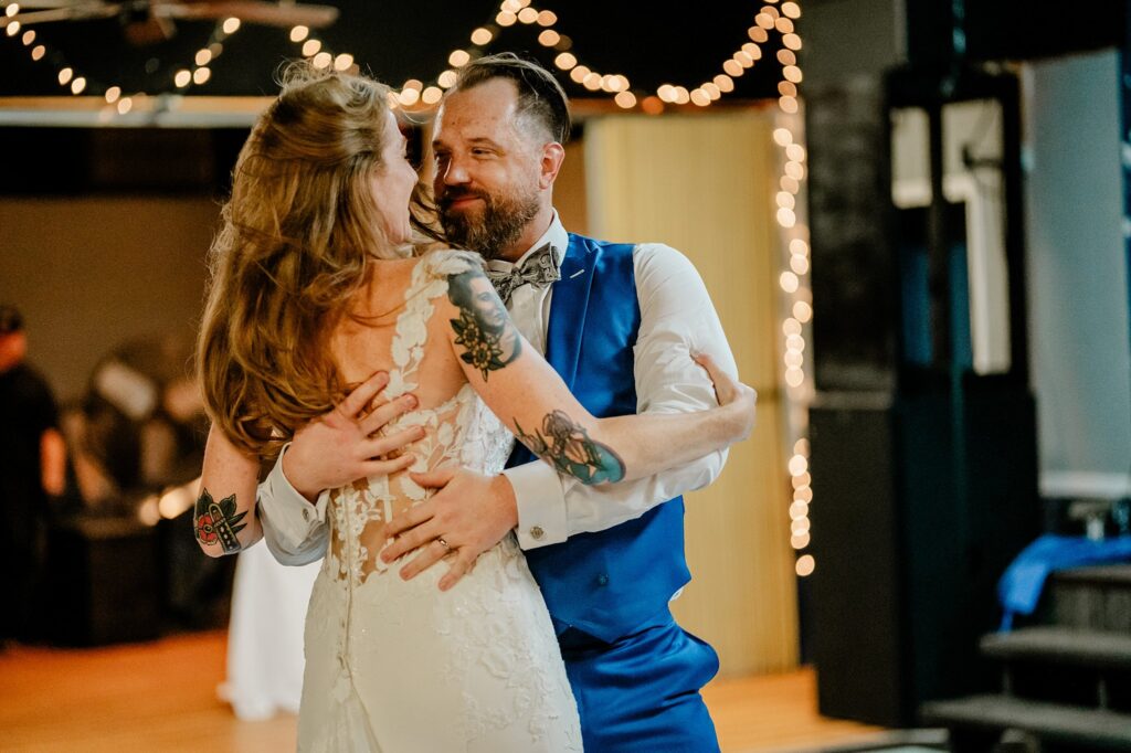 A bride and groom embrace and smile while dancing, surrounded by soft, warm string lights in a decorated indoor venue. Captured by a Nashville wedding photographer, the bride wears a lace gown and the groom is in a blue vest and bow tie.