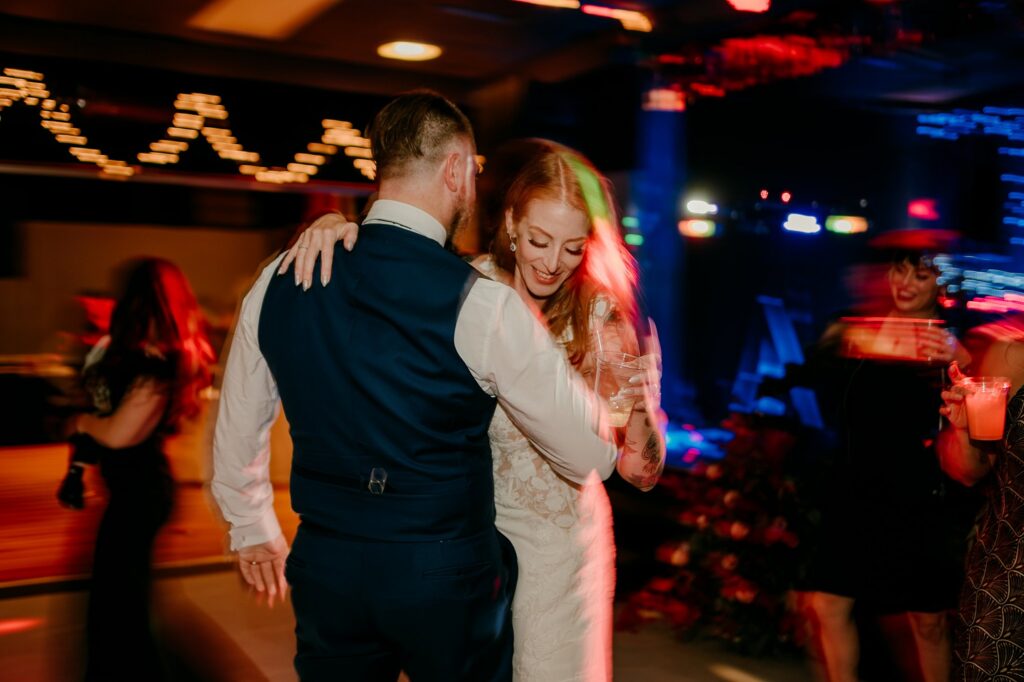 A couple dances closely at a lively event, with the woman smiling in a white dress and the man in a suit vest. Captured by a Nashville wedding photographer, colorful, blurred lights and guests add to the festive, joyful atmosphere.