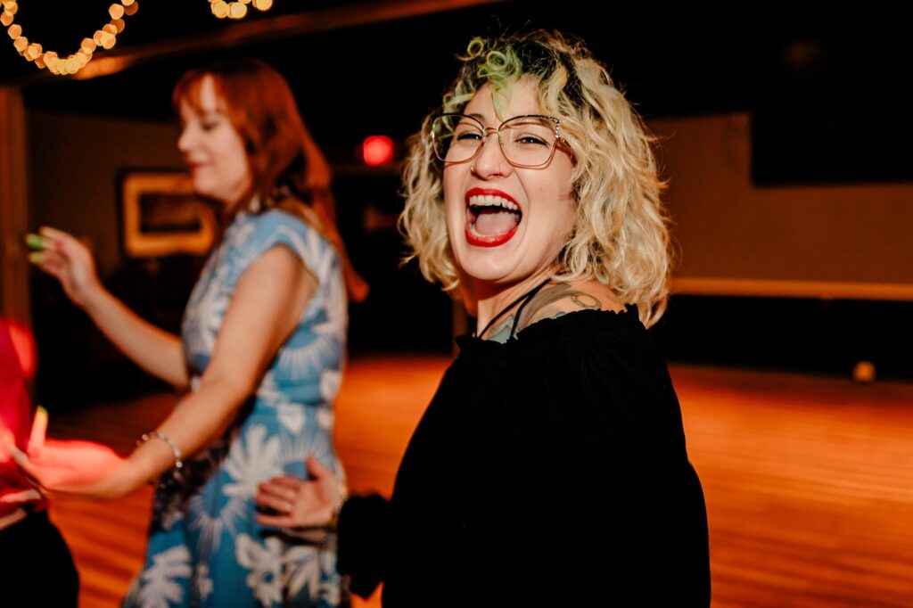 A woman with curly blonde hair, glasses, and red lipstick smiles and laughs at the camera while dancing indoors. Captured by a Nashville wedding photographer, another woman in a blue dress with white patterns dances in the warm, festive lighting.