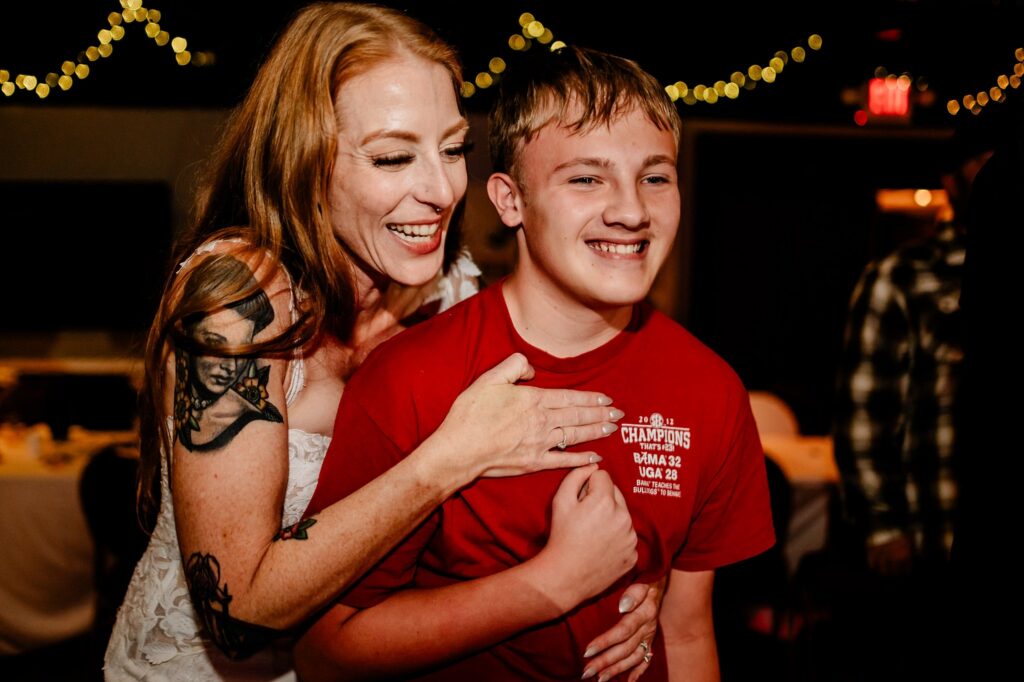 A woman with long red hair and tattoos joyfully hugs a smiling boy in a red shirt at an indoor event with string lights, perfectly captured by a Nashville wedding photographer.