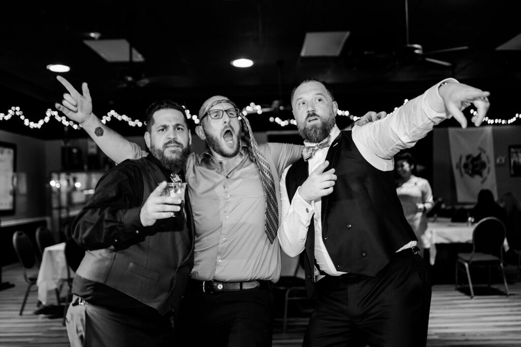 Three men in dress clothes pose energetically at an indoor event, one holding a drink, another with his mouth open in excitement, and the third pointing forward. String lights decorate the ceiling—a lively moment captured by a Nashville wedding photographer.
