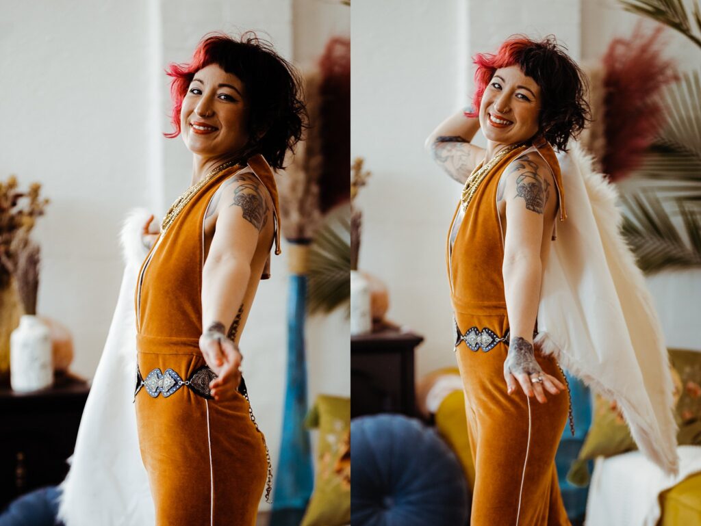 A smiling person with short dark hair and a red streak poses playfully in a mustard yellow jumpsuit and feathered cape. Captured by a Nashville creative portrait photographer, they stand in a warmly lit, stylish room filled with plants and decor.