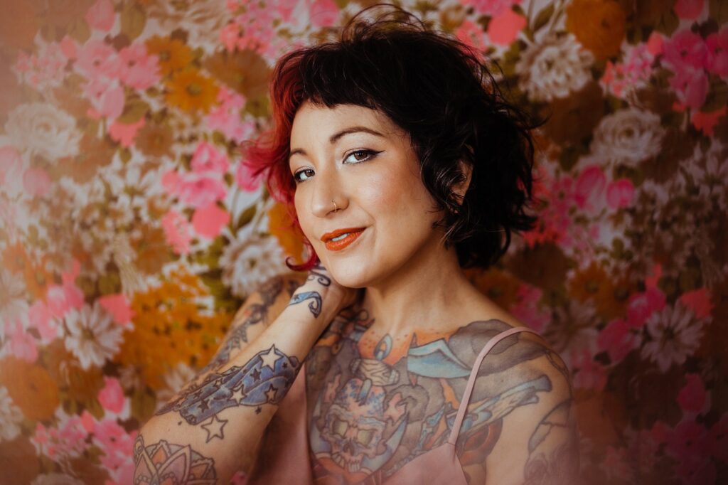 A person with short dark hair, tattoos on their arms and chest, and orange lipstick smiles softly in front of vibrant floral wallpaper—an image captured by a Nashville creative portrait photographer.