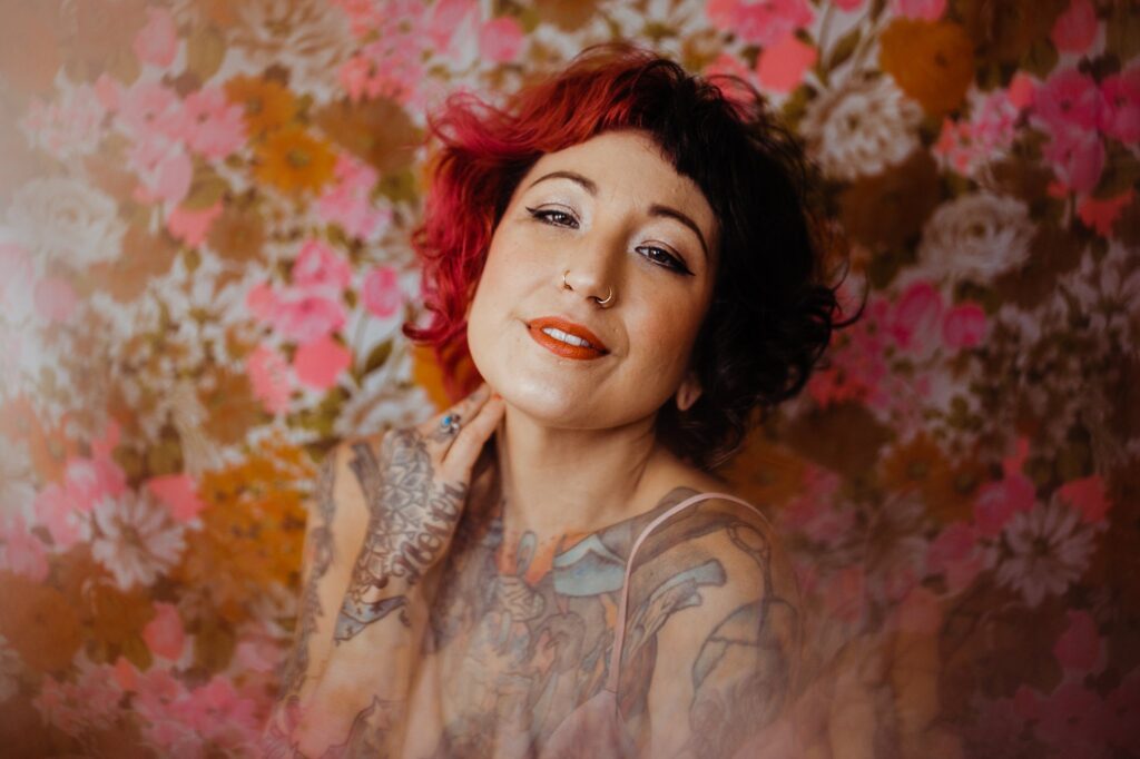 A person with short, curly, dark and red hair, wearing orange lipstick and a nose ring, poses smiling in front of a vibrant floral background. Captured by a Nashville creative portrait photographer, their arm and chest tattoos are on display.