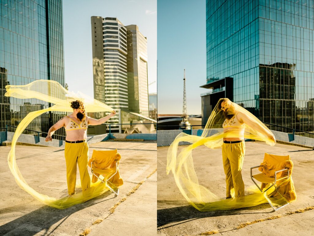 A person in yellow pants and a beige top stands on a rooftop, waving flowing yellow fabric near a chair. Tall glass buildings reflect sunlight in the background, captured by a Nashville creative portrait photographer.