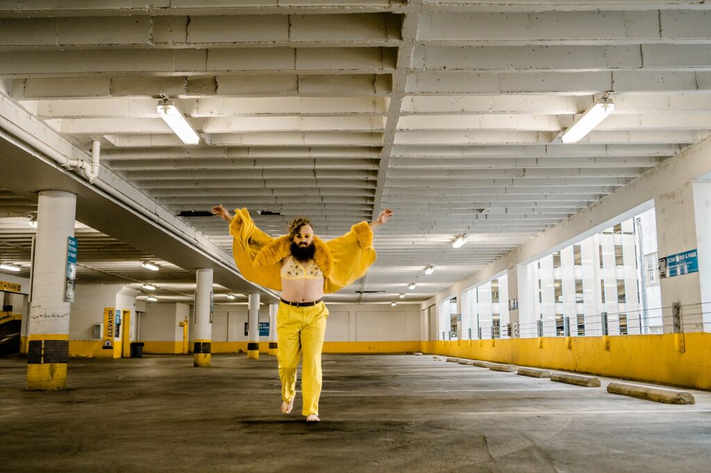 A person in a yellow outfit and crop top dances barefoot in an empty parking garage with yellow and white walls, arms outstretched and smiling. Bright ceiling lights illuminate the scene—captured by a Nashville creative portrait photographer.