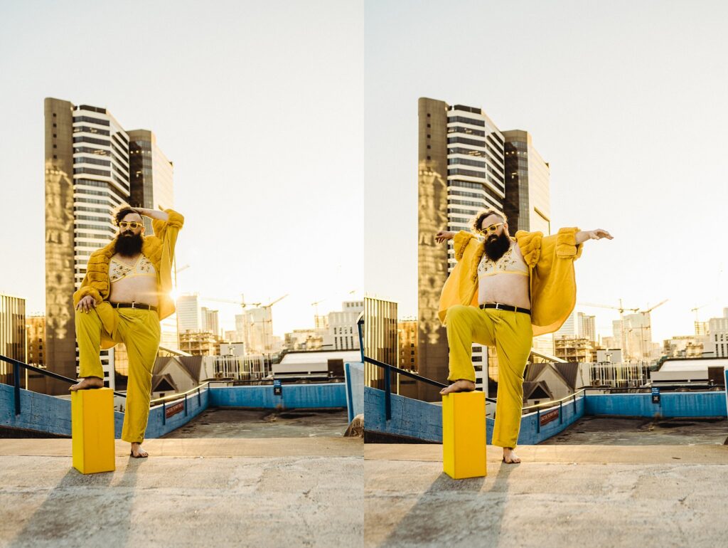 A bearded person in yellow pants, a yellow jacket, and sunglasses poses energetically on a rooftop at sunset with tall buildings behind them and a yellow suitcase by their feet—perfect inspiration from a Nashville creative portrait photographer.