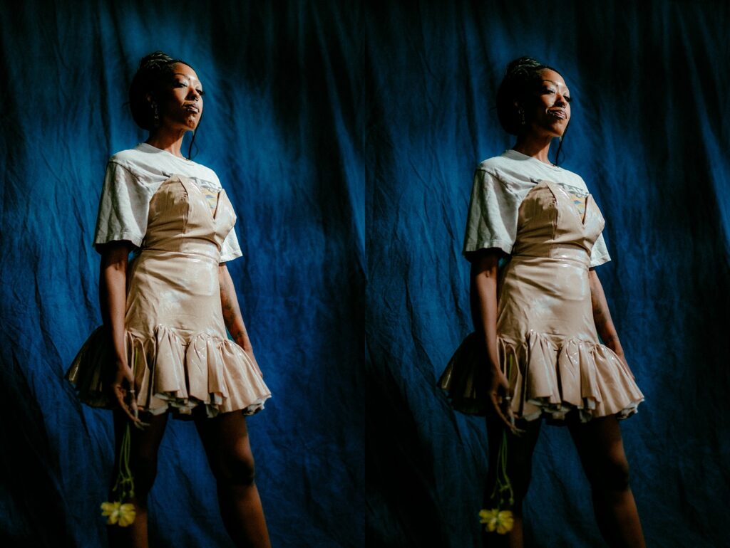 A woman in a shiny beige dress layered over a white T-shirt stands against a dark blue backdrop, holding a yellow flower. Captured by a Nashville creative portrait photographer, the duplicated image highlights her calm, confident expression.