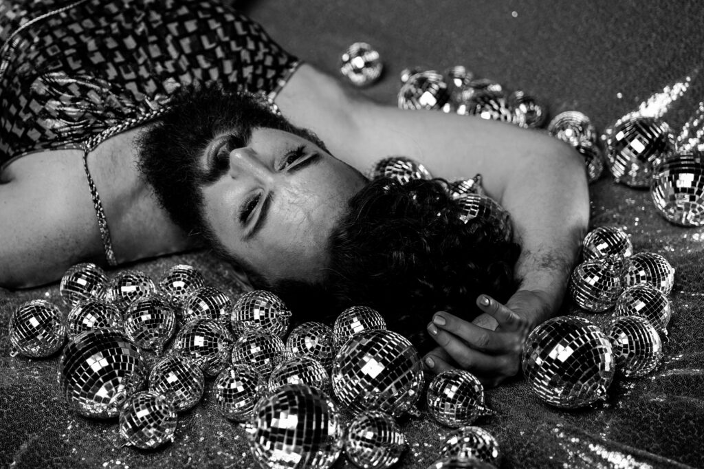 A bearded person in a patterned dress lies on a glittery surface, surrounded by small disco balls, with one hand resting behind their head and eyes closed. Captured by a Nashville creative portrait photographer in striking black and white.
