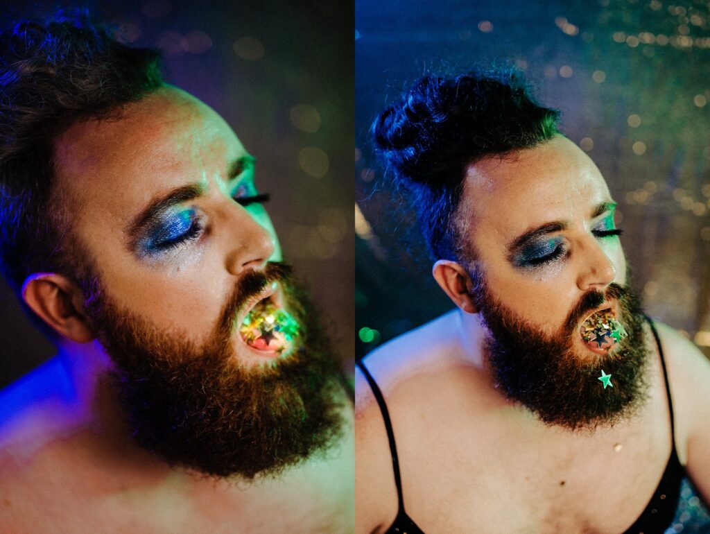 A bearded person with glittery blue eyeshadow and dramatic makeup poses with their mouth open, revealing colorful, shiny decorations inside. Captured by a Nashville creative portrait photographer, they wear a black top and a star sticker on their chin against a sparkly background.