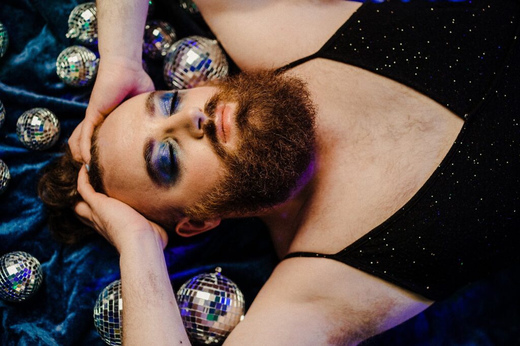 A person with a beard and bold blue eye makeup lies on blue fabric, wearing a black sparkly top, surrounded by small disco balls. Captured by a Nashville creative portrait photographer, their hands rest on their head in striking style.