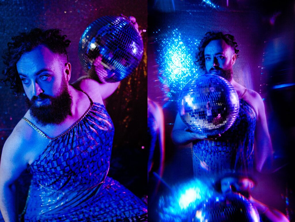 A person with a beard and curly hair poses with a disco ball, wearing a shimmery dress. Captured by a Nashville creative portrait photographer, vibrant blue and purple lights set a dramatic, festive atmosphere.