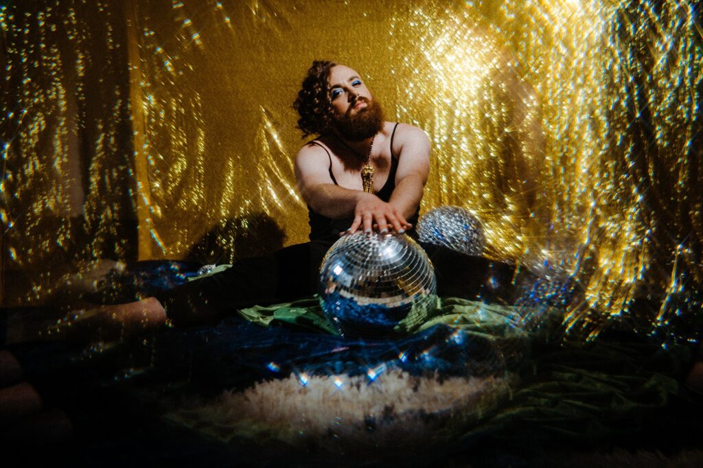 A person with curly red hair and a beard sits on the floor, surrounded by disco balls and shimmering gold fabric, as sparkling light reflections create a dreamy, festive atmosphere—captured by a Nashville creative portrait photographer.