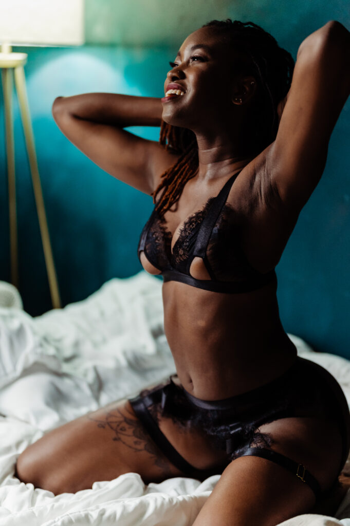 A woman in black lingerie sits on a bed with white sheets, smiling and stretching her arms behind her head. Exuding boudoir elegance, she radiates confidence while a lamp stands in the background against a teal wall.