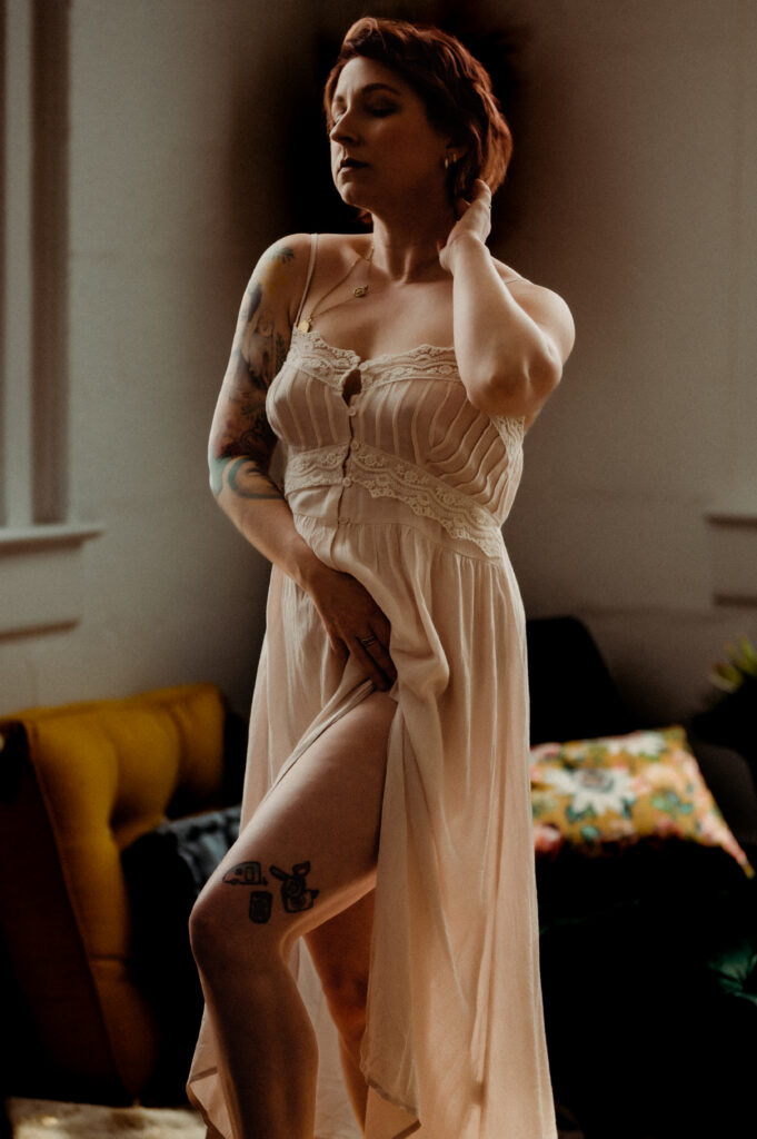A person with short hair and visible tattoos poses in a softly lit room, wearing a light, lacy nightgown and touching their neck and thigh, looking sexy with one leg exposed through a high slit—a perfect boudoir photography moment.