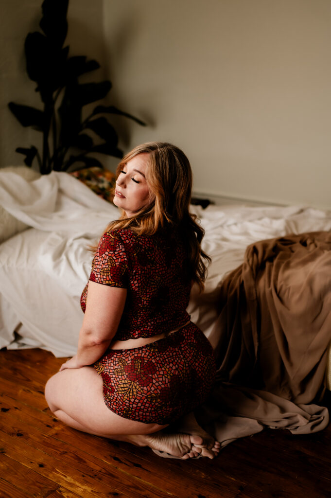 A woman with blonde hair kneels on a wooden floor beside an unmade bed, wearing a short-sleeve, patterned outfit. Soft natural light and neutral-toned sheets create a boudoir atmosphere as she poses, looking sexy and serene.