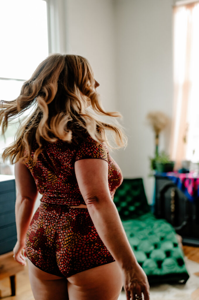A woman with wavy blonde hair wearing a colorful patterned romper stands indoors, mid-turn, with blurred motion suggesting movement. The soft natural light and her confident pose evoke a boudoir photography vibe.