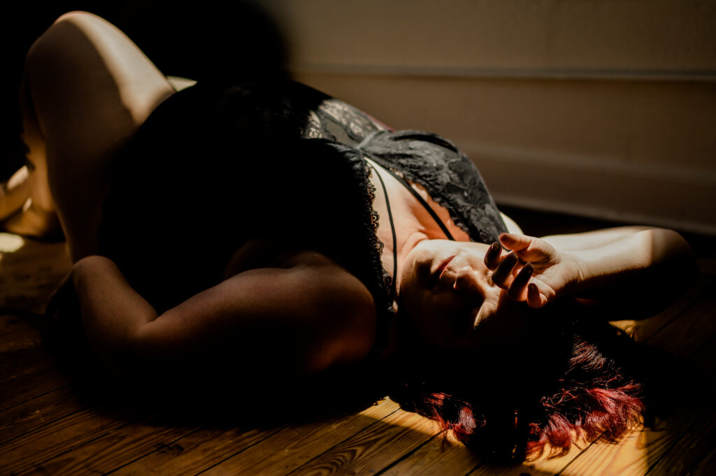 A person in black lace lingerie lies on a wooden floor, partially in shadow and sunlight, with one arm resting on their forehead and eyes closed. The softly lit scene captures a boudoir moment that is intimate and subtly looking sexy.